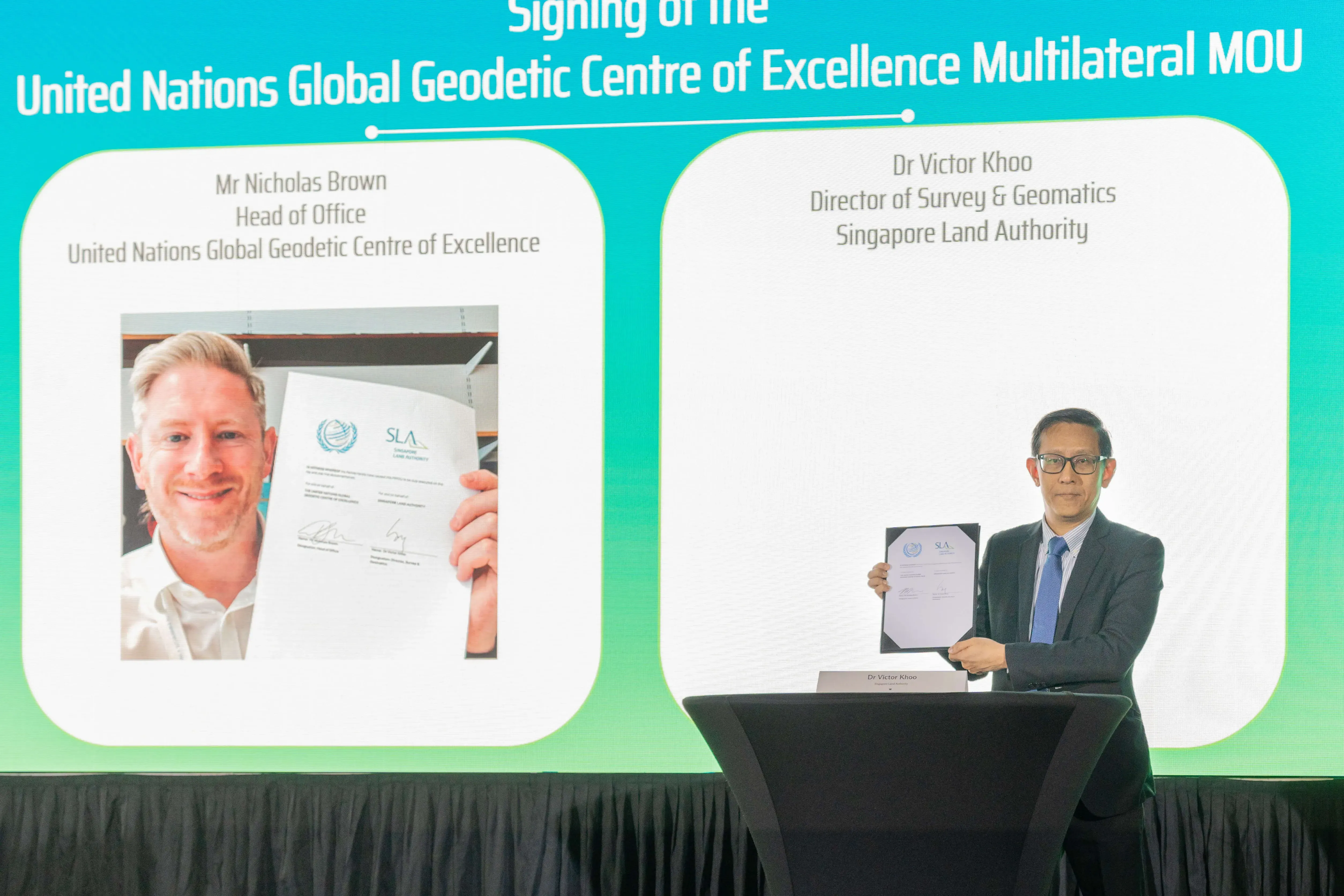 Dr Victor Khoo holds a signed MOU document at a podium beside a screen showing Mr Nicholas Brown at the UN MOU signing.